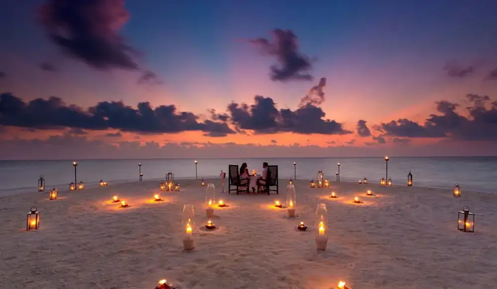 A candlelit table for two on the sand at sunset, the lagoon beyond