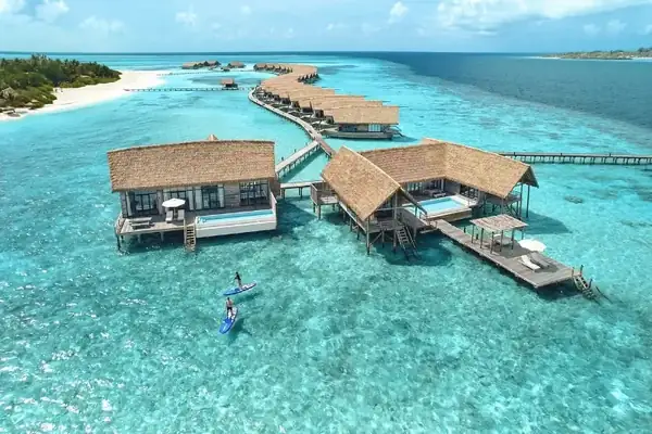 Overwater villas above clear shallow water, with paddleboarders in the lagoon