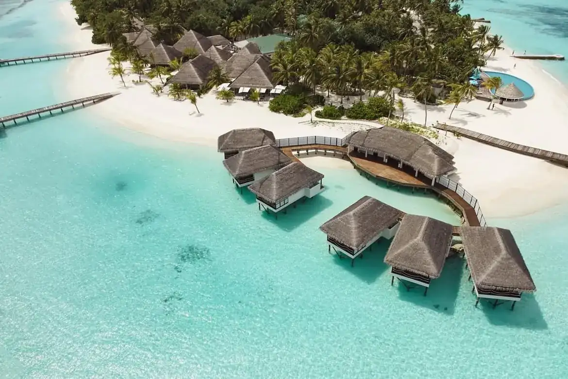 An aerial view of a Maldives island: thatched overwater villas curving out from a palm-covered beach into a pale turquoise lagoon