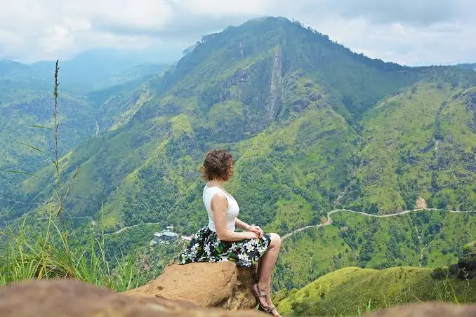 Woman sitting on a rock overlooking green mountains and a winding road in Sri Lanka's hill country
