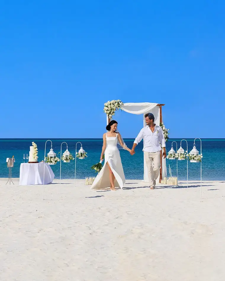 A couple walking hand in hand from a flower-draped arch on white sand